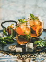 Summer refreshing cold peach ice tea with fresh mint in glass jars on metal tray over oriental ceramic tile background, selective focus, copy space