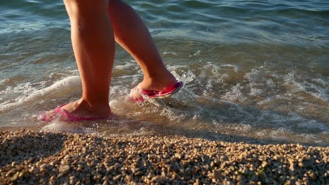 girl walking in pink sandals on sand shore with waves splashing her feets in slow motion hd