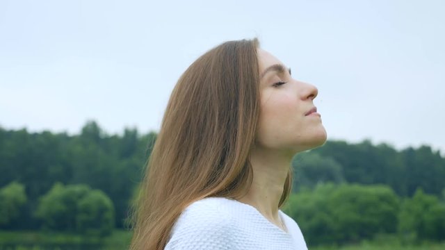 Caucasian girl deeply breathe with closed eyes on the nature background, her hair flying in the wind, then she slowly turns and looks into the camera. 4K video of relaxation and stress relief.