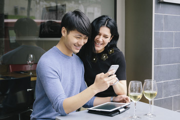 Multiethnic couple in a restaurant looking at phone with a glass of white wine