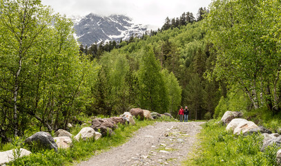 A couple walks along a mountain road through a forest in the background of snow mountains