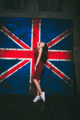 красивая девушка с темными волосами в коротком, бордовом платье и белых кедах возле британского флага