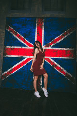 Obraz na płótnie Canvas красивая девушка с темными волосами в коротком, бордовом платье и белых кедах возле британского флага