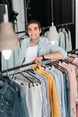 handsome young boutique owner leaning at hangers with clothes and smiling at camera