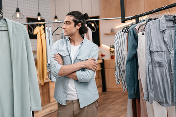 handsome young man standing between hangers with clothes and looking away in boutique