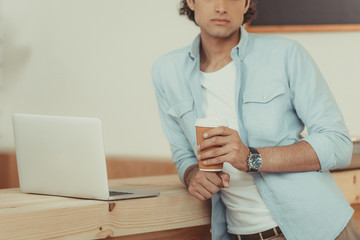 cropped shot of young man using laptop and drinking coffee in paper cup while working