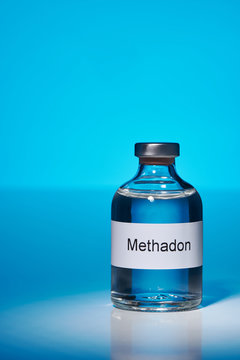 Eine Flasche mit Methadon steht im Spot licht auf einer weißen Fläche vor einem blau leuchtenden Hintergrund