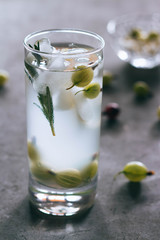 Lemonade with ice in a glass cup, opposite light, from lemon, gooseberry, rosemary, on a concrete background