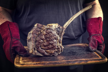 Man is holding barbecue wagyu tomahawk steak on old burnt cutting board in his hands with gloves
