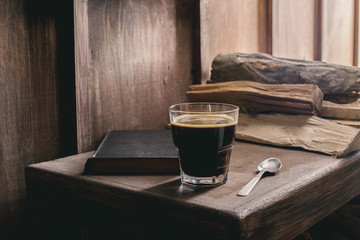 Glass of coffee drink, old book and spoon on rustic wooden table near pile of firewood