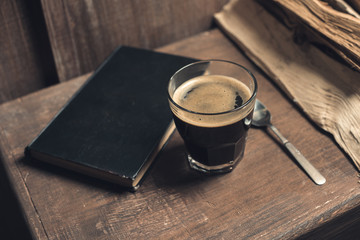 Glass of coffee drink, old book and spoon on rustic wooden table near pile of firewood