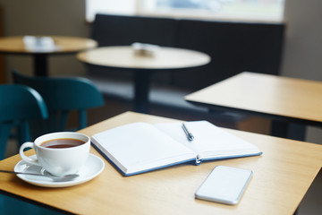 Open notebook with pen, cup of tea and smartphone on wooden table in cafe