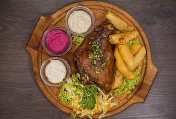 Fried pork leg drumstick, knee of a wild boar with vegetables of red and yellow tomatoes, garlic, parsley and basil and potatoes on a wooden background