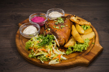 Fried pork leg drumstick, knee of a wild boar with vegetables of red and yellow tomatoes, garlic, parsley and basil and potatoes on a wooden background