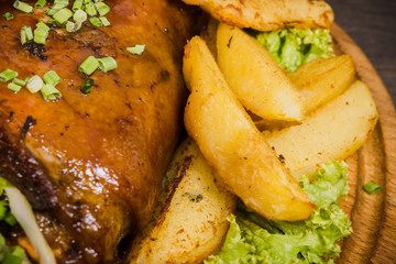 Fried pork leg drumstick, knee of a wild boar with vegetables of red and yellow tomatoes, garlic, parsley and basil and potatoes on a wooden background