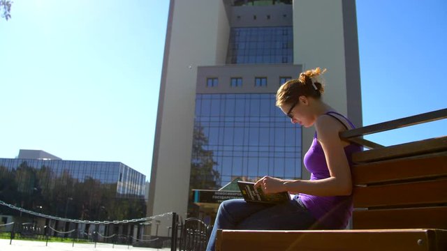 The girl is sitting on a bench in front of the university and reading a book