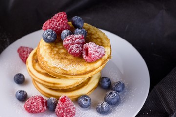 Stack of pancakes with raspberries and blueberries  coated with powdered sugar on white plate and black background
