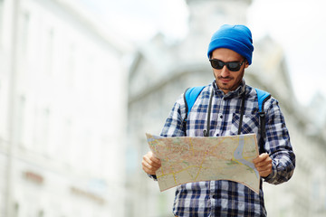 Portrait of contemporary young man on solo trip in Europe, standing in street of old city looking at map tourist guide