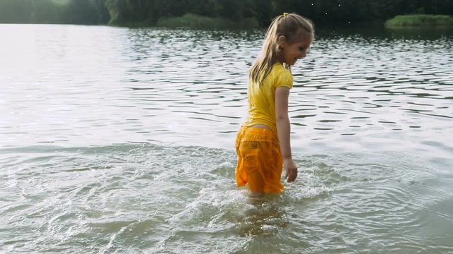 Little girl in yellow wet clothes runs and splashes happily in the water of the forest lake. Slow motion.
