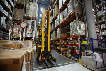 Yellow forklift truck waiting in aisle between rows of tall racks with packed goods in empty warehouse
