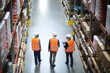 Group of warehouse workers wearing hardhats and reflective jackets waking in aisle between tall...