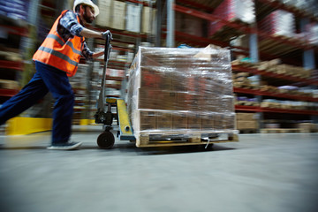 Blurred motion shot of warehouse worker wearing hardhat and reflective jacket pushing moving cart with boxes along isle between tall racks - obrazy, fototapety, plakaty