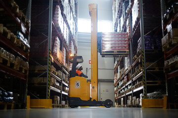 Side view  portrait of warehouse worker using reach fork truck to load pallet with boxes on tall rack