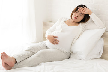Asian pregnant woman with hand on forehead suffering headache sitting on bed at home