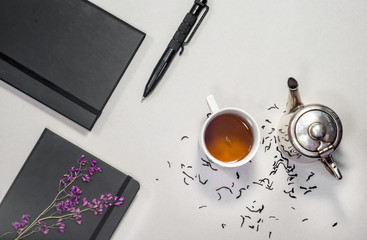 Silver teapot, white vintage cup, notebooks, pen, spoon, dry tea leafs, handle on gray background. Business concept