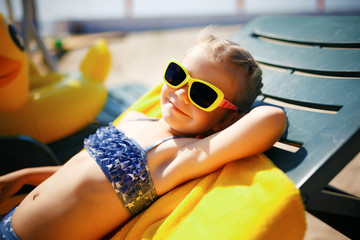 little girl in sunglasses is sunbathing on a sunbed near the pool at the resort