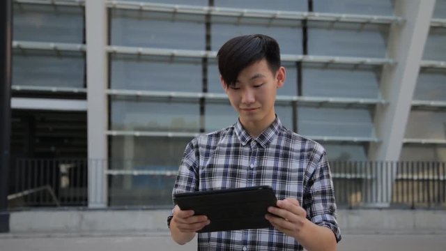 Asian man in city using tablet computer PC outdoors wearing checkerboard shirt.
