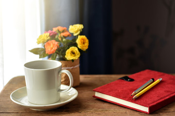 White cup of hot drinks on wooden table by the window. Small vibrant color roses and red cover notebook with pencils on it as background. Hot coffee or tea for a cold morning refreshment.