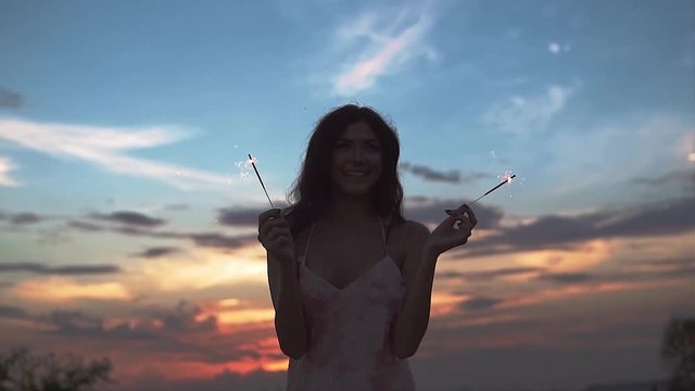 sparklers in the hands of happy girl against the sky at sunset. portrait of a beautiful girl in a cocktail dress. slow motion
