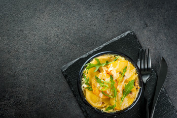 Obraz na płótnie Canvas Casserole. French cuisine. Homemade potato gratin in a bowl for baking. On a black stone background. With leaves of fresh arugula for decoration. Top view copy space