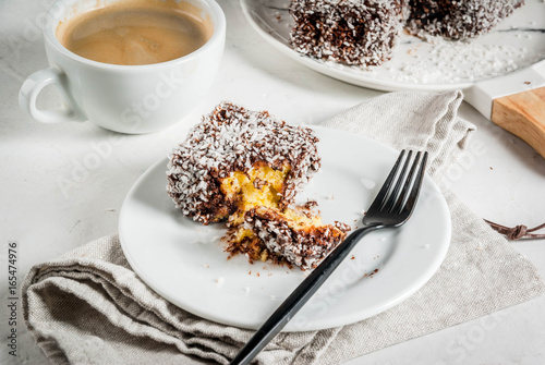 Australian food. Traditional dessert Lamington - pieces of biscuit in dark chocolate, sprinkled with coconut powder chips. On a marble plate, white table. With coffee mug. Copy space