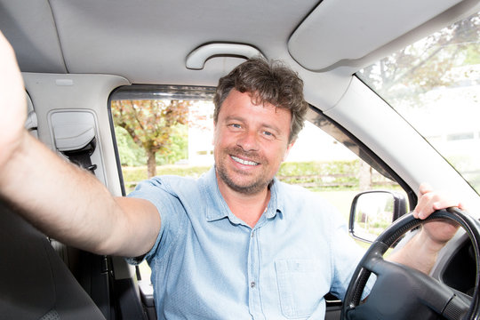 Enjoying his road trip. Cheerful man smiling at camera and make smartphone selfie sitting inside of his minivan