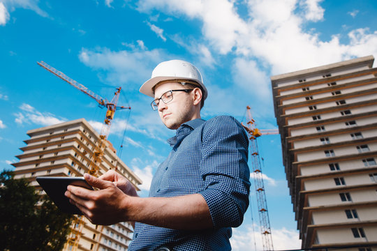 builder is a man in a protective helmet, glasses and a tablet computer in the hands of a crane, a multi-storey house and a blue sky. Concept construction, business, technology