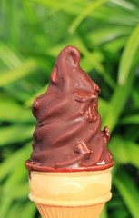 Closed up chocolate dipped soft serve ice cream cone, with blurred green bush in background 