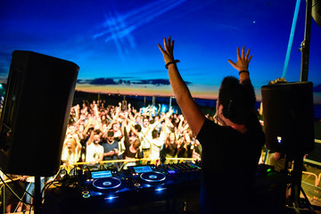 Dj and dance floor with hands to the sky during a beach party at sunrise. Emotions for the sound explosion at music festival. The view is from behind the consolle on the stage - obrazy, fototapety, plakaty