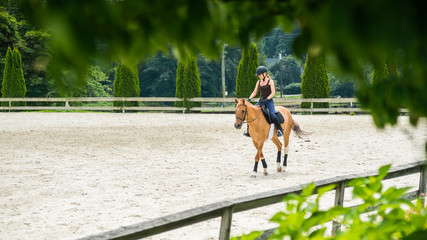 woman horseback riding beautiful brown mare and training in sandy outdoors manege at horse ranch.