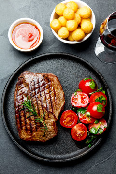 Grilled beef steak served on cast iron plate with tomato salad, potatoes balls and red wine. Barbecue, bbq meat beef tenderloin. Top view, slate background