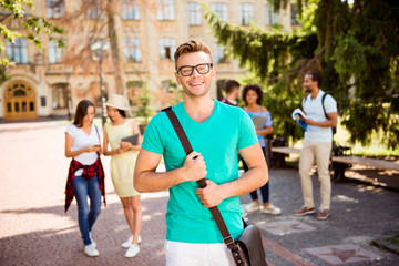 Young successful blond nerdy student is standing with bag and smiles, behind are his classmates, park near campus, sunny day, carefree and enjoyable mood