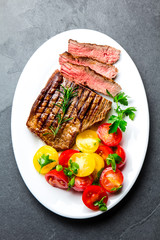Sliced medium rare grilled beef steak served on white plate with tomato salad and potatoes balls. Barbecue, bbq meat beef tenderloin. Top view, slate background, copy space