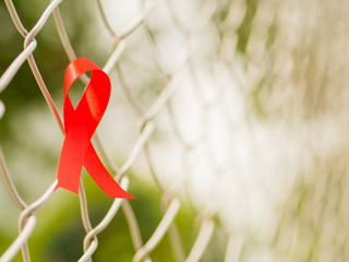 World AIDS day concept. Red ribbon hang on metal net and tree leaves bokeh background.