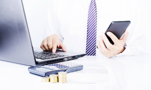 Business man touching cellphone and typing laptop with calculator and coins stack on office desk for finance ideas concept