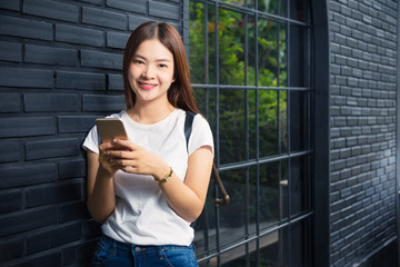 young happy woman in white tshirt and blue jeans standing leaning against dark brick wall using her cell phone, concept of young hipster generation lifestyle using technology