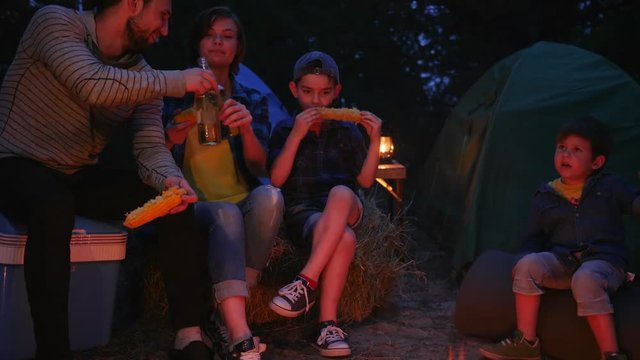 parent and children enjoying camping in woodland on vacation, happy family eating fried corn and drinking beverage near fire into forest, mom and dad with sons resting at night near tent in camp,