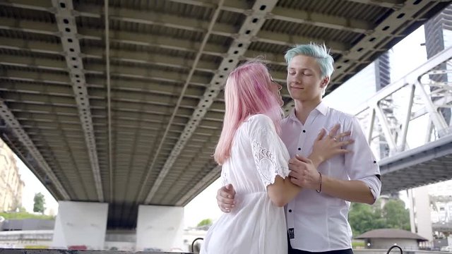 young couple in white shirts meet under a highway bridge across the river to a large modern city. a man with blue hair and girl with pink. they are a modern, informal people