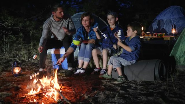 happy family fry marshmallow at fire in travel camp in forest, mom and daddy with kids resting to campground and roast zephyr on bonfire, parents with childs broil marshmallow on campfire to woodland,