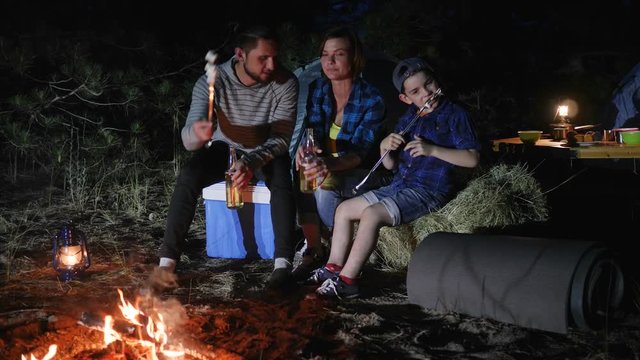 family trip on weekend in forest, travel camp of american family in woodland, parents drink beer and child eats zephyr near bonfire in journey with tents, mom and dad with son enjoy camping on nature,